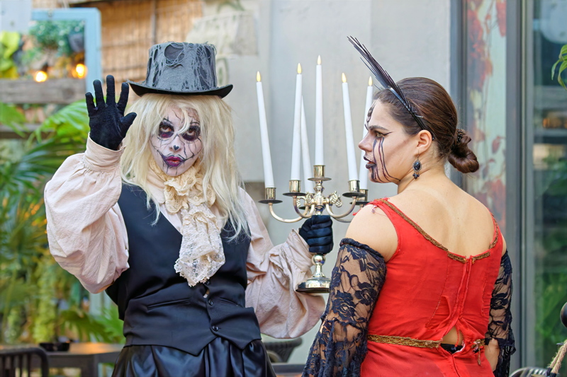 13 - Women dressed in Halloween costumes, with one person wearing a top hat and pale face paint while holding a candelabrum, and the other person wearing a red dress with dark makeup